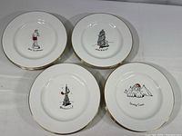 Four different designs of white porcelain plates with gold rim accents arranged in square layout showing Christmas themed illustrations and phrases.