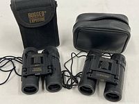 Pair of compact binoculars side-by-side with their respective belt pouches: Rugged Exposure with canvas pouch, Simmons with leatherette pouch.