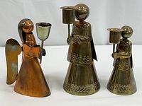 Three vintage angel candlestick holders; two brass and one copper, standing upright with candle cups.