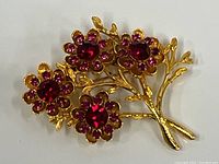 Close-up front view of the gold-tone flower motif brooch with red glass/crystal stones arranged as flower petals with a larger stone at the center of each flower.