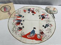 Large round silk table cover with hand painted Japanese women figures and floral border, showing some age spots and minor paint cracking.