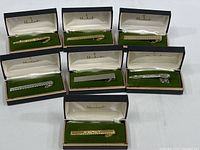 Seven Marbert men's tie clips displayed in original boxes, four gold-toned and three silver-toned, each about 2 inches long.