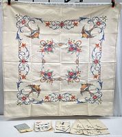 Wide view of the cream-colored linen tablecloth with colorful floral embroidery in cross-stitch surrounded by a blue scroll border. Below, six embroidered napkins and an additional set of various napkins and towel packages are displayed.