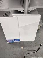 Two white blank canvases, one smaller and one larger, standing against shelf with visible wiring on floor.