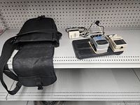 Image showing the black carrying case with flap open, two pedals, and power adapter on grey shelf.