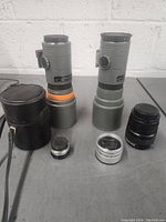 Overview of six camera lenses including two large gray telephoto lenses, a black Olympus lens, and three smaller vintage style lenses, as well as a leather carrying case for one telephoto lens.