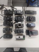 Full top-down photo showing all 13 cameras and a case on white table surface.