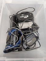 Lot of four older handheld video cameras in a plastic bin, tangled with cables and carrying straps. Shows three Sony brand camcorders and one silver Sharp camcorder.