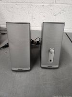 Front view of the pair of Bose silver speakers on a gray surface against a white textured wall.