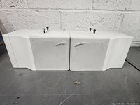 Front view of two white Bose speakers positioned side by side on a gray concrete surface against a brick wall background.