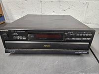 Front and top view of Panasonic 5-disc CD changer showing brand, control buttons, digital display and disc tray.