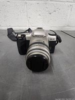 Front view of the silver and black Pentax ZX-30 camera with attached zoom lens and lens cap.