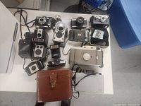 Wide view of thirteen vintage cameras arranged on a table including box, rangefinder, and toy cameras along with flash units and carrying cases.