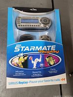 Front view of Sirius Starmate Replay satellite radio in its original blue packaging showing the display unit and remote control inside the box.
