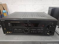 Front view of black Sony STR-DE315 Digital Audio Video Control Center/Receiver showing front panel display, buttons and ports on grey surface with power cable connected.