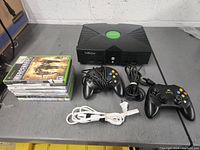 Original Xbox console on table with three controllers, six game cases, and bundled power cable.