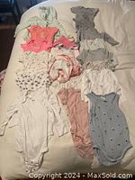 Photo shows 16 baby girl clothing pieces including bodysuits and outerwear in pastel colors laid out on a bed.