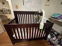 Solid dark brown wood crib with vertical slats and a baby mobile attached on one side. Mattress covered with pink and white striped fabric and fitted sheet, with some baby toys inside.