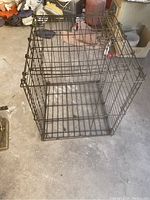 Single photo showing a metal wire dog crate standing on a floor in a garage or storage area, viewed from above at an angle, giving a clear view of the crate structure and door latch.