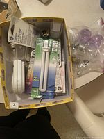 Box containing assorted light bulbs including CFL tubes in packaging, loose incandescent bulbs, and some lighting accessories.