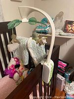 White baby crib mobile with green accents and four plush toys hanging, mounted on side of crib next to stuffed animals and other baby items.