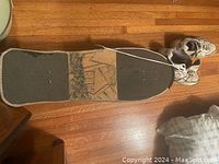 Photo showing wooden skateboard deck with grip tape and worn design, placed on wooden floor
