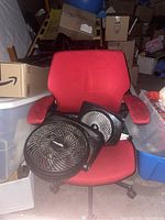 Red fabric office chair with a black Vornado fan placed on the seat, surrounded by boxes and storage bins.