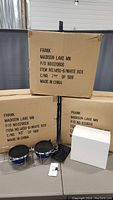 Three shipping boxes labeled Frank, Madison Lake MN, containing travel pet bowls. Displayed on table are two shiny blue pet bowls in chrome metal stands with black lids.