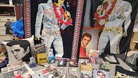 Photo showing the full range of Elvis Presley collectible items including tapestries, pillows, books, CDs, DVDs, novelty money, and small collectibles