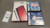 Red Nintendo DS Lite handheld console box, two Nintendo DS game cases (Big Brain Academy and Brain Age 2) on gray surface