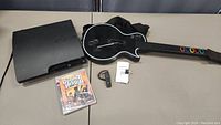 Full view of PlayStation 3 console, Guitar Hero guitar controller, and factory sealed Guitar Hero III Legends of Rock game case on a flat surface.