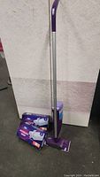 Full view of Swiffer Wet Jet mop, two boxes of cleaning pads, and new bottle of cleaner on floor against a light wall.