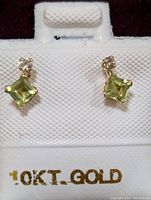 Pair of yellow gold earrings each with a square-cut light green peridot and a small round diamond above it, displayed on white leather padding stamped '10KT GOLD'.
