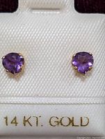 Pair of heart-shaped purple amethyst earrings set in 14K yellow gold, displayed on a white textured holder marked '14 KT. GOLD'.