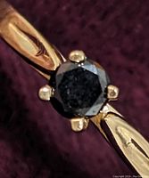 Close-up photo of the round faceted black diamond set in a 10K yellow gold four-prong setting ring on a dark fabric background.