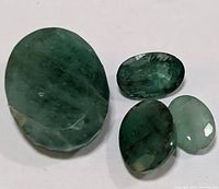 Four genuine faceted emerald gemstones displayed on white background, varying sizes and shades of green visible. Oval and rounded shapes. Emerald clarity typical with inclusions visible.
