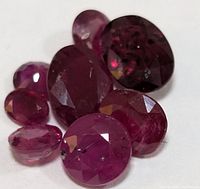 Seven faceted ruby gemstones arranged on a white background showing various sizes and cuts with deep pink to reddish hues and visible inclusions.