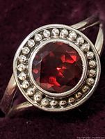 Close-up photo of sterling silver ring with a central round faceted garnet gemstone surrounded by silver bead accents, against a dark red fabric background.
