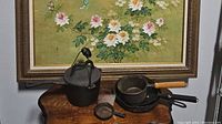 Five cast iron cookware pieces on wooden table under floral framed painting. Includes various saucepans, skillets, and griddle.