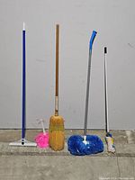Five cleaning tools lined up against a wall: a small carpet rake, a pink feather duster, a straw broom, a blue multi surface brush, and a white cotton mop handle.