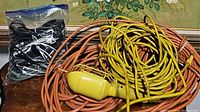 Yellow extension cord with construction light, long brown extension cord, and bag of assorted smaller cords on wood surface
