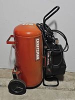 Orange Craftsman vertical air compressor tank with attached black motor and handle, wheels included for mobility.