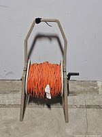 Front view of beige plastic hose reel cart with bright orange extension cord wrapped around, showing handle and crank mechanism.