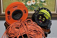 Three items on a table in front of floral framed artwork: an orange cord reel, a gray Masterplug cord reel with green cord, and a loose orange extension cord.