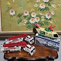 Two Hess fire trucks and Hess helicopter on original box displayed on wood surface with floral painting background.