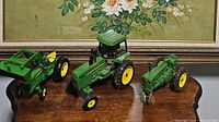 Four vintage metal John Deere toy tractors and farming equipment in green and yellow colors arranged on wooden table with floral painting in background