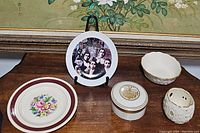 Five porcelain items arranged on a wood surface including two plates, a vial or box with lid, an egg-shaped holder, and a scalloped bowl.