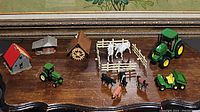 Photo shows the farm animals including white horses, black cow, pig, brown animals, green tractors with yellow wheels, trailers, and several small farm buildings with red and gray roofs.