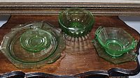 Lot with five vintage green depression glass pieces displayed on wood surface beneath framed artwork. Items include ornate plate, engraved saucer, block optic bowl, handled bowl, and handled platter.