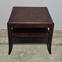 Front view of the walnut brown side table with one shelf, showing the slight curve in legs and storage shelf beneath the tabletop.